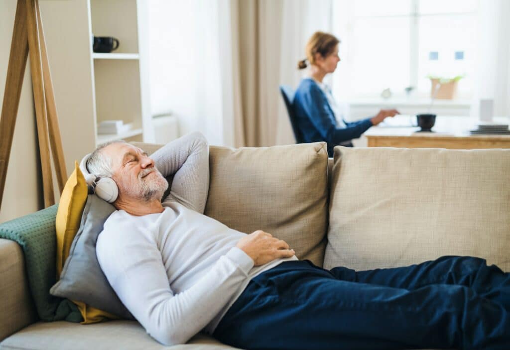 A happy senior couple indoors with a pet dog at home, using laptop and headphones.